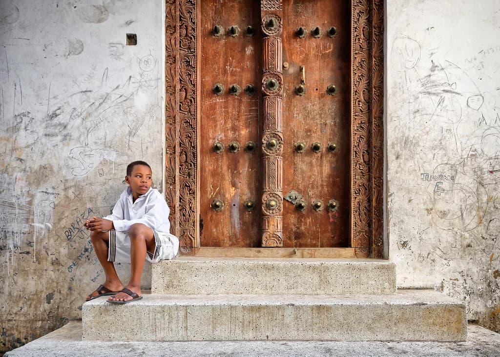 Un ragazzo seduto sui gradini di una porta in legno intagliato a Stone Town, Zanzibar
